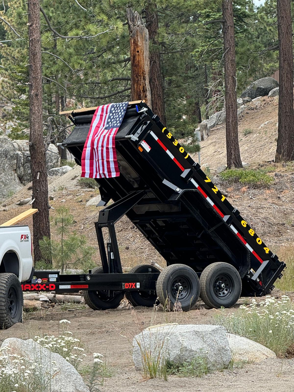 Stamparini Services black MAXX-D dump trailer tilted with an American flag, parked in the Tahoe pines — phone (530) 721-6804 painted on the side